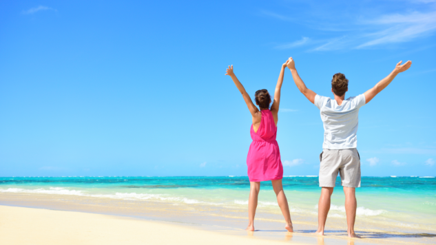 Couple standing on a sandy beach in the sun, holding hands with their arms raised in the air