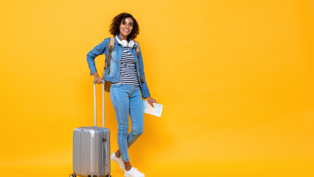 Woman standing with a suitcase and holding her passport against a yellow background