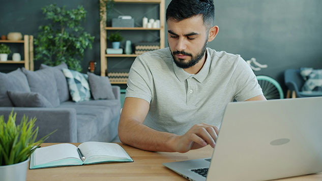 Man using a laptop at a table with an open notebook beside him.